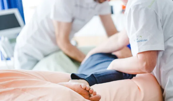 Zwei Personen in weißen Arbeitskleidung führen eine Behandlung an einem Patienten durch, der auf einer Liege mit einem rosa Laken liegt. Ein Fuß des Patienten ist sichtbar. Im Hintergrund sind medizinische Geräte und ein Vorhang zu sehen.