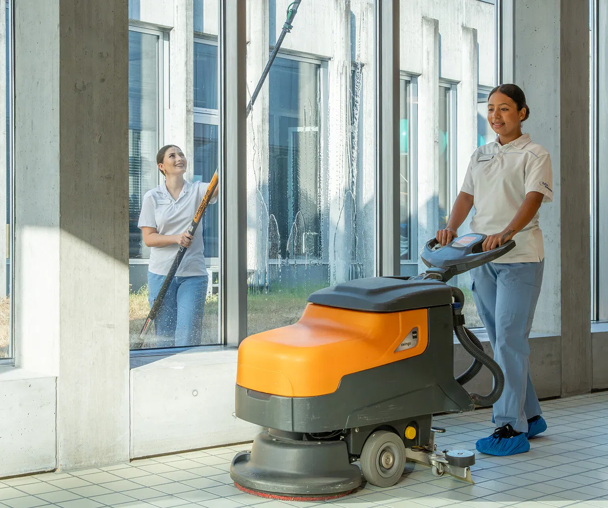 Zwei Reinigungskräfte reinigen einen hellen Flur mit großen Fenstern: Eine bedient eine orange-graue Scheuersaugmaschine, die andere wischt die Fenster.