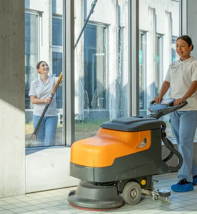 Zwei Reinigungskräfte reinigen einen hellen Flur mit großen Fenstern: Eine bedient eine orange-graue Scheuersaugmaschine, die andere wischt die Fenster.