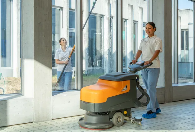 Zwei Reinigungskräfte reinigen einen hellen Flur mit großen Fenstern: Eine bedient eine orange-graue Scheuersaugmaschine, die andere wischt die Fenster.