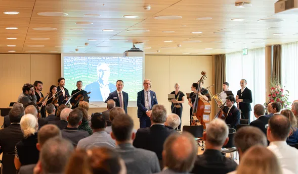 Publikum bei einer Veranstaltung mit kleinem Orchester, zwei Rednern und projizierter Präsentation im Hintergrund.