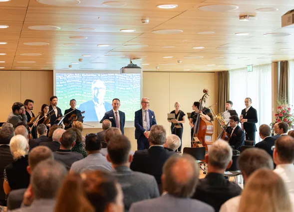 Publikum bei einer Veranstaltung mit kleinem Orchester, zwei Rednern und projizierter Präsentation im Hintergrund.