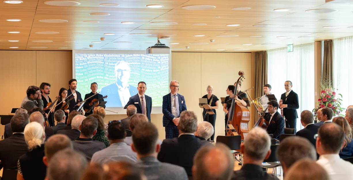 Publikum bei einer Veranstaltung mit kleinem Orchester, zwei Rednern und projizierter Präsentation im Hintergrund.