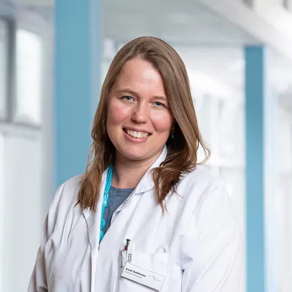 Lächelnde Ärztin mit langen braunen Haaren im weißen Kittel und Namensschild vor hellem Klinikinterieur.
