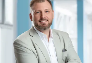 Mann mit kurzem Bart und lächelndem Gesicht, trägt einen hellen Blazer über einem weißen Hemd, steht mit verschränkten Armen vor einem hellen, modernen Hintergrund.