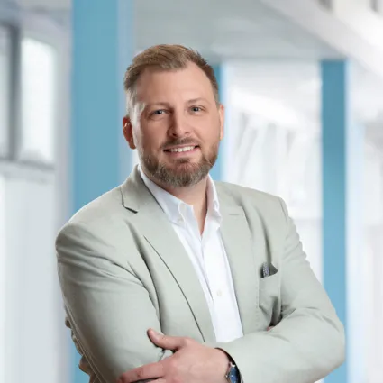 Mann mit kurzem Bart und lächelndem Gesicht, trägt einen hellen Blazer über einem weißen Hemd, steht mit verschränkten Armen vor einem hellen, modernen Hintergrund.