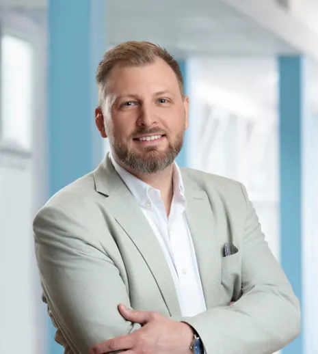 Mann mit kurzem Bart und lächelndem Gesicht, trägt einen hellen Blazer über einem weißen Hemd, steht mit verschränkten Armen vor einem hellen, modernen Hintergrund.