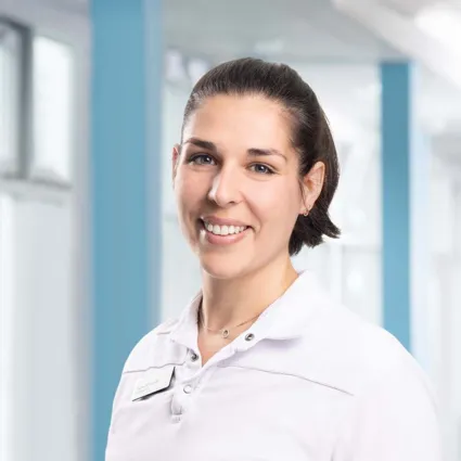 Lächelnde Frau mit kurzen, dunklen Haaren, trägt ein weißes Poloshirt und eine Namensschilder, vor einem hellen, modernen Hintergrund mit blauen Akzenten.