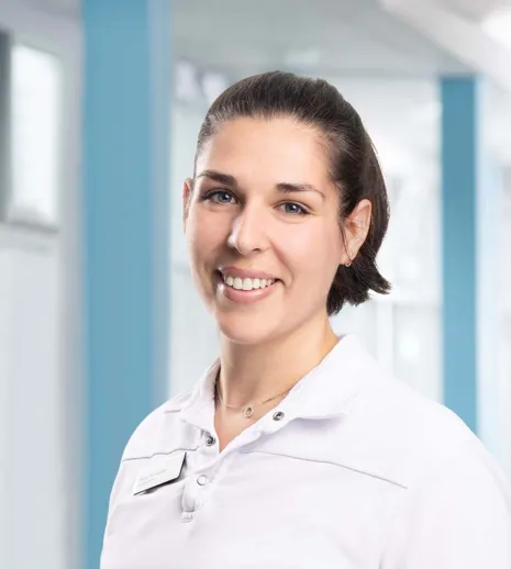 Lächelnde Frau mit kurzen, dunklen Haaren, trägt ein weißes Poloshirt und eine Namensschilder, vor einem hellen, modernen Hintergrund mit blauen Akzenten.