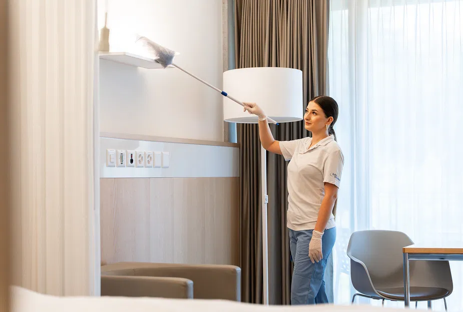 Pflegerin mit Handschuhen wischt in einem hellen Patientenzimmer mit einem Staubwedel ein Wandregal ab. Mobiliar, Stehlampe und Vorhänge im Hintergrund.