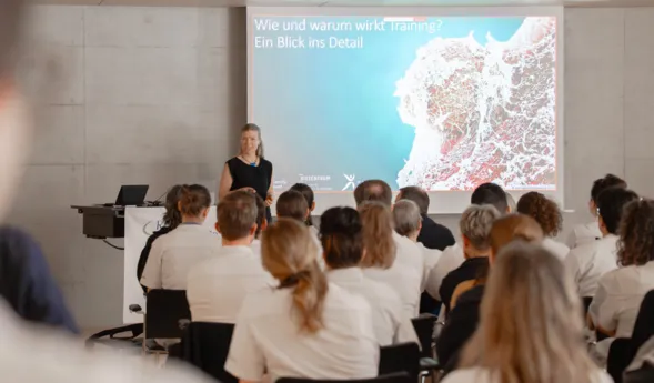 Vortragsraum mit Publikum in Stuhlreihen: Eine Referentin steht am Rednerpult und präsentiert eine Folie mit dem Titel „Wie und warum wirkt Training?“ und einer Karte vor großer Leinwand.