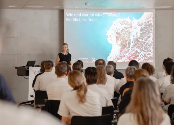 Vortragsraum mit Publikum in Stuhlreihen: Eine Referentin steht am Rednerpult und präsentiert eine Folie mit dem Titel „Wie und warum wirkt Training?“ und einer Karte vor großer Leinwand.