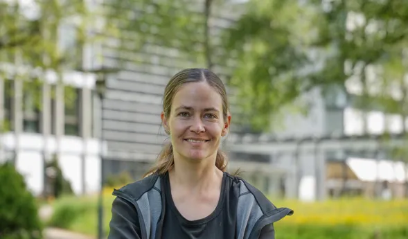 Lächelnde Frau mit Fahrradhelm ohne Helm, sitzt/steht auf einem Fahrrad in einem grünen Parkweg; unscharfer Hintergrund mit Bäumen und Gebäudefassade.