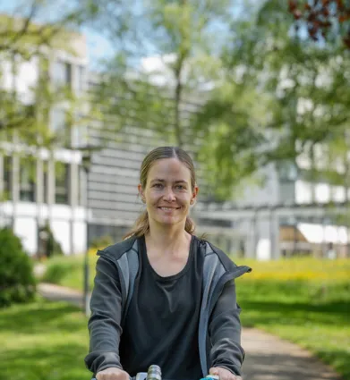 Lächelnde Frau mit Fahrradhelm ohne Helm, sitzt/steht auf einem Fahrrad in einem grünen Parkweg; unscharfer Hintergrund mit Bäumen und Gebäudefassade.