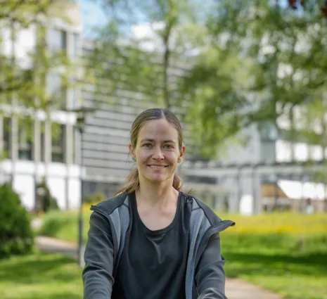 Lächelnde Frau mit Fahrradhelm ohne Helm, sitzt/steht auf einem Fahrrad in einem grünen Parkweg; unscharfer Hintergrund mit Bäumen und Gebäudefassade.