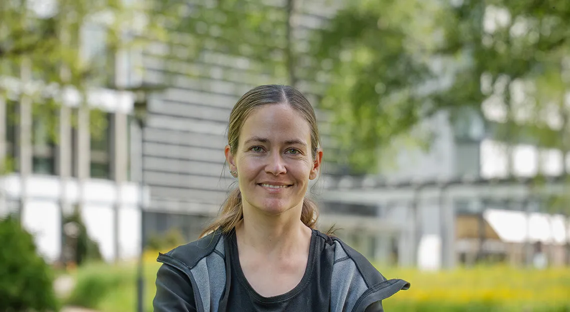 Lächelnde Frau mit Fahrradhelm ohne Helm, sitzt/steht auf einem Fahrrad in einem grünen Parkweg; unscharfer Hintergrund mit Bäumen und Gebäudefassade.