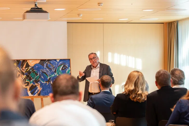 Mann hält einen Vortrag vor sitzendem Publikum in einem hellen Konferenzraum mit Leinwand und abstraktem Gemälde.