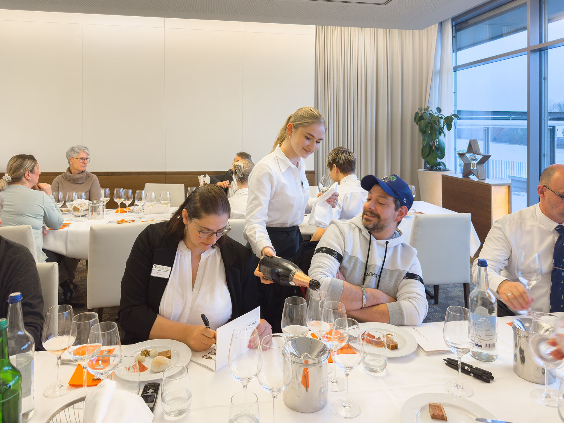 Elegante Restaurantatmosphäre mit mehreren Gästen an einem Tisch. Eine Kellnerin serviert Sekt an einen Mann, der in einem grauen Sweatshirt sitzt. Eine Frau in Schwarz schreibt an einem Tisch, während andere Gäste im Hintergrund miteinander sprechen....