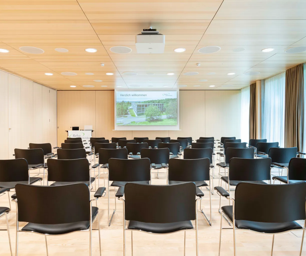 Konferenzraum mit Reihen von schwarzen Stühlen, einem Projektor an der Decke und einer Leinwand, auf der 