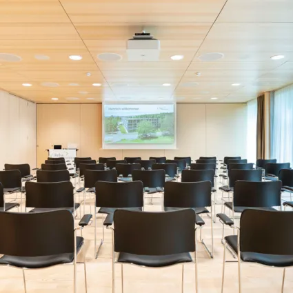 Konferenzraum mit Reihen von schwarzen Stühlen, einem Projektor an der Decke und einer Leinwand, auf der 