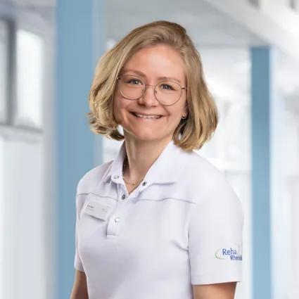 Porträt einer freundlichen Frau mit kurz blondem Haar und Brille im weißen Firmen-Poloshirt vor hellem blau-weißen Hintergrund.