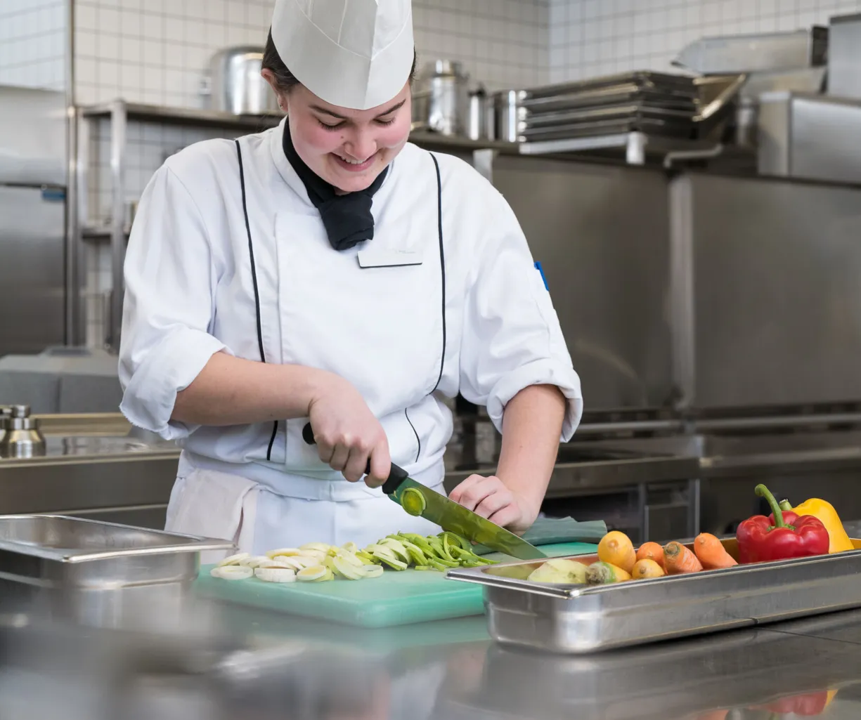 Köchin in einer modernen Küche schneidet frisches Gemüse auf einem grünen Schneidebrett. Neben ihr liegen bunte Paprika, Karotten und andere Zutaten in Edelstahlbehältern.