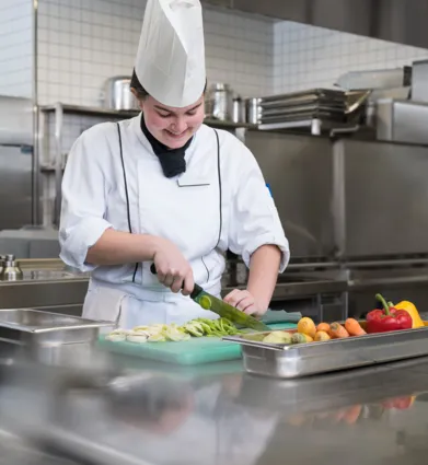 Köchin in einer modernen Küche schneidet frisches Gemüse auf einem grünen Schneidebrett. Neben ihr liegen bunte Paprika, Karotten und andere Zutaten in Edelstahlbehältern.
