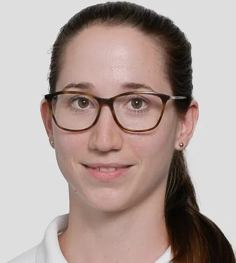Porträt einer lächelnden Frau mit Brille, zurückgebundenem braunem Haar und weißem Kragen vor grauem Hintergrund.
