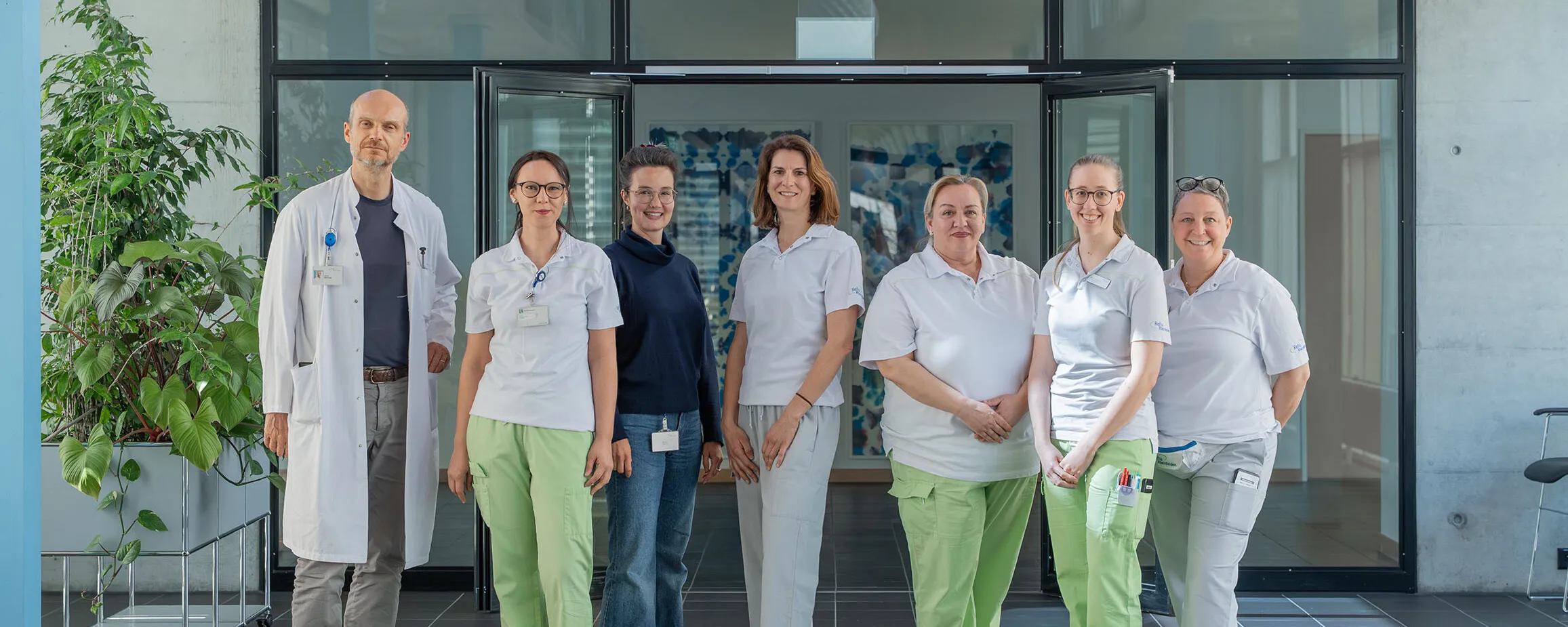 Team aus mehreren Ärztinnen und einem Arzt in weißen Kitteln und grünen Hosen vor einer Glasfront, im Eingangsbereich eines modernen Gebäudes.