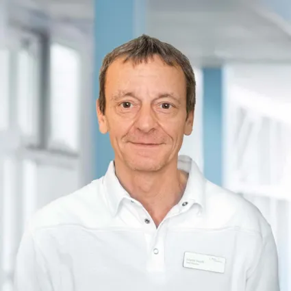 Mann mit kurzen, braunen Haaren und leichtem Bart, trägt ein weißes Polohemd. Im Hintergrund sind unscharfe, helle Wände und Fenster zu sehen. Ein Namensschild mit der Aufschrift 