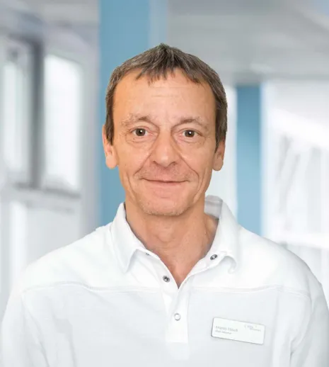 Mann mit kurzen, braunen Haaren und leichtem Bart, trägt ein weißes Polohemd. Im Hintergrund sind unscharfe, helle Wände und Fenster zu sehen. Ein Namensschild mit der Aufschrift 