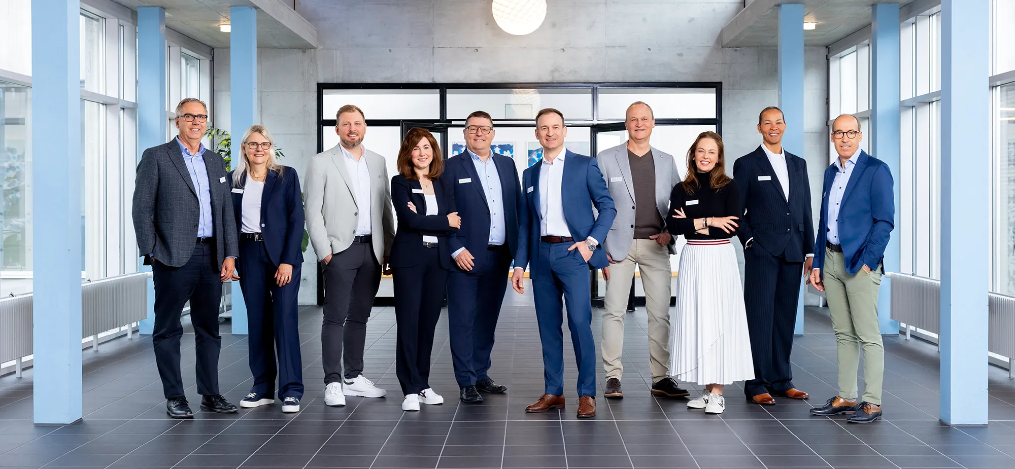 Gruppenszene mit zehn Personen in formeller Kleidung, die in einem modernen Raum mit großen Fenstern und grauem Boden stehen. Einige Personen lächeln, während andere neutral blicken. Im Hintergrund sind Pflanzen und eine große Glaswand sichtbar.