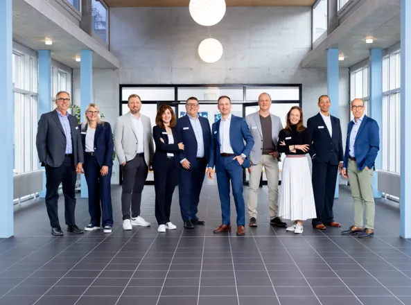 Gruppenszene mit zehn Personen in formeller Kleidung, die in einem modernen Raum mit großen Fenstern und grauem Boden stehen. Einige Personen lächeln, während andere neutral blicken. Im Hintergrund sind Pflanzen und eine große Glaswand sichtbar.