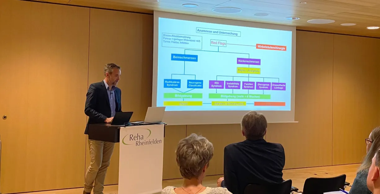 Präsentation in einem Seminarraum mit einem Referenten, der an einem Laptop steht. Im Hintergrund ist eine Folie mit einem Diagramm zu Anamnese und Untersuchung bei Beinschmerzen und Rückenschmerzen zu sehen. Teilnehmende sitzen im Vordergrund, einige...