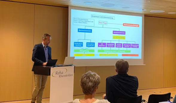 Präsentation in einem Seminarraum mit einem Referenten, der an einem Laptop steht. Im Hintergrund ist eine Folie mit einem Diagramm zu Anamnese und Untersuchung bei Beinschmerzen und Rückenschmerzen zu sehen. Teilnehmende sitzen im Vordergrund, einige...