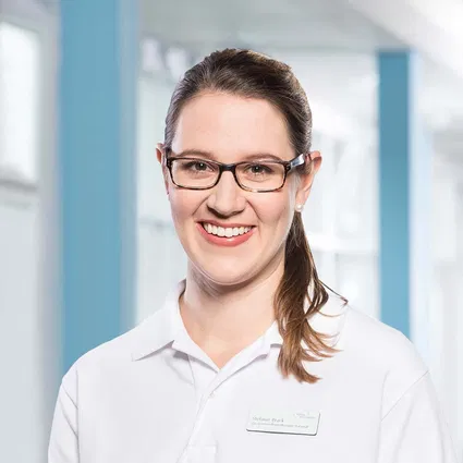 Lächelnde Frau mit Brille und weißem Poloshirt, Namensschild, vor hellem blau-weißem Hintergrund.