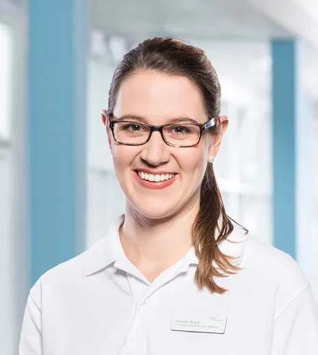 Lächelnde Frau mit Brille und weißem Poloshirt, Namensschild, vor hellem blau-weißem Hintergrund.