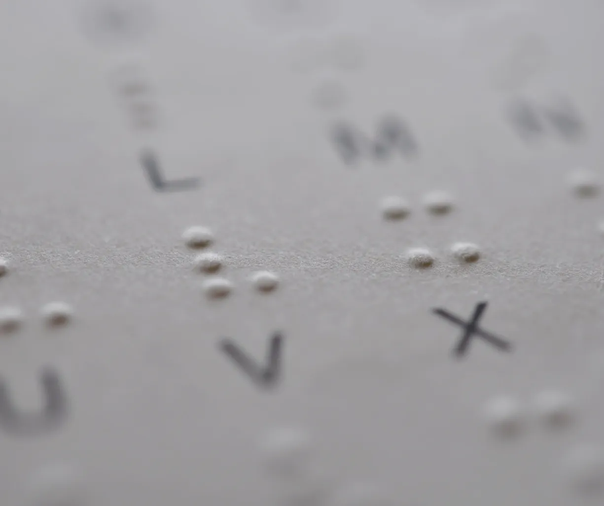 Braille-Schrift auf einem Blatt Papier mit erhabenen Punkten und gedruckten Buchstaben, darunter die Buchstaben K, L, M, N, U, V und X.
