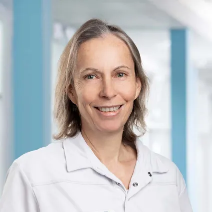 Lächelnde Frau mit kurzen, grauen Haaren in einem weißen Oberteil, vor einem hellen, modernen Hintergrund.