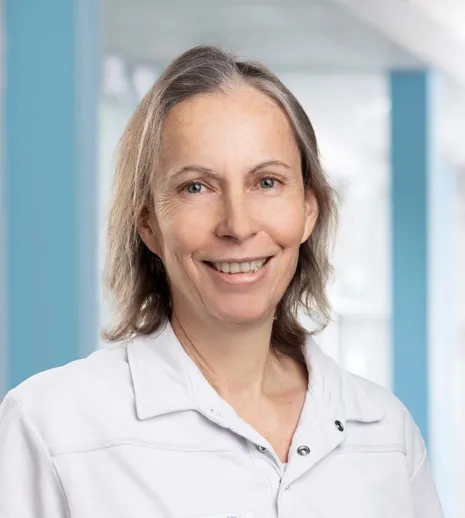 Lächelnde Frau mit kurzen, grauen Haaren in einem weißen Oberteil, vor einem hellen, modernen Hintergrund.