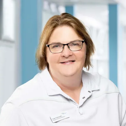 Frau mit kurzen, blonden Haaren und Brille, trägt ein weißes Oberteil mit einem Kragen. Im Hintergrund sind helle Wände und Fenster zu sehen.