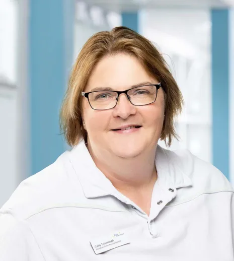 Frau mit kurzen, blonden Haaren und Brille, trägt ein weißes Oberteil mit einem Kragen. Im Hintergrund sind helle Wände und Fenster zu sehen.