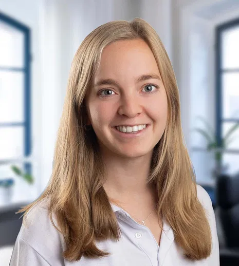 Lächelnde junge Frau mit langen blonden Haaren und weißer Bluse vor hellem Bürohintergrund.