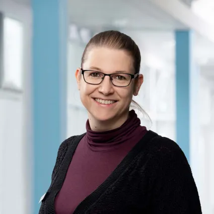 Frau mit Brille, lächelnd, trägt einen lila Rollkragenpullover und eine schwarze Strickjacke, vor einem hellen, modernen Hintergrund mit blauen Akzenten.