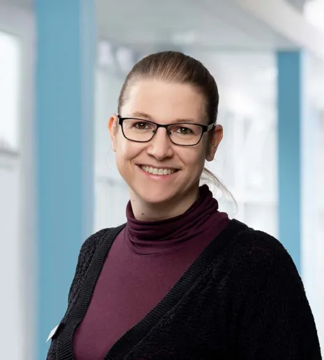 Frau mit Brille, lächelnd, trägt einen lila Rollkragenpullover und eine schwarze Strickjacke, vor einem hellen, modernen Hintergrund mit blauen Akzenten.