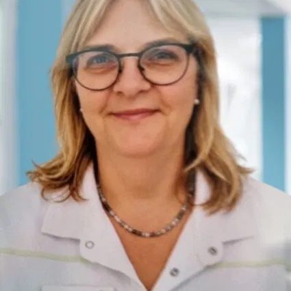 Porträt einer lächelnden Frau mit blondem Haar und Brille in weißem Arbeitskittel mit Namensschild und Halskette.
