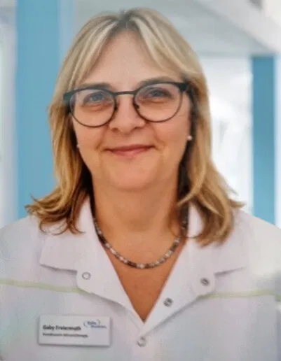 Porträt einer lächelnden Frau mit blondem Haar und Brille in weißem Arbeitskittel mit Namensschild und Halskette.