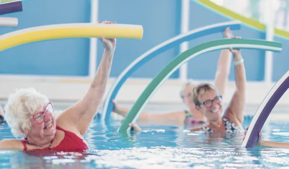 Ältere Frauen im Wasser mit bunten Schwimmnudeln, die Übungen machen. Helle, freundliche Umgebung mit blauen und grünen Wänden. Einige Frauen lächeln, während sie aktiv sind. Reflektionen auf der Wasseroberfläche sind sichtbar.