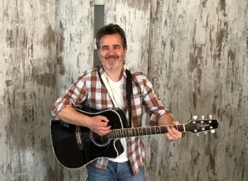Lächelnder Mann mit kariertem Hemd spielt eine schwarze Akustikgitarre vor rustikaler Holzwand.