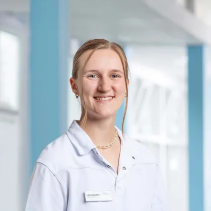 Porträt einer lächelnden Frau in weißem Arztkittel mit Namensschild vor hellem, blauem Klinikflur.